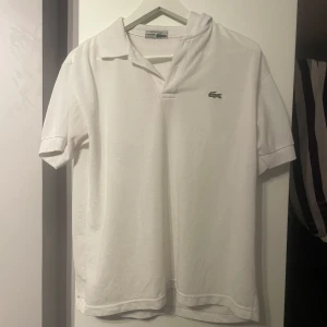Vit pikétröja från Lacoste - Säljer en klassisk vit pikétröja från Lacoste med den ikoniska krokodilloggan på bröstet. Tröjan har korta ärmar och en stilren krage. Perfekt för en avslappnad och stilren look.
