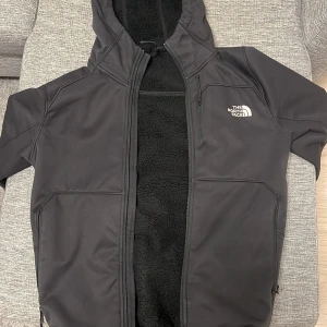 Svart jacka från The North Face - Säljer en svart jacka från The North Face med huva och dragkedja. Jackan har en stilren design med logotyp på bröstet och ryggen. Perfekt för kyligare dagar.