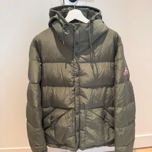 Grön dunjacka från Holubar - Säljer en grön dunjacka från Holubar med huva och broderad logga på ärmen. Jackan har en quiltad design och stängs med dragkedja och knappar. Perfekt för kyliga dagar. Endast använd ett fåtal gånger. Nypris 6700 kr