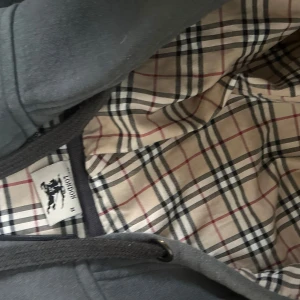 Grå huvtröja med rutigt foder - Säljer en snygg grå burberry huvtröja med dragkedja och ett klassiskt rutigt foder. Tröjan har ribbade muddar och små broderade detaljer på bröstet. Perfekt för höst och vår även vinter , både stilren och bekväm. Passar till vardags och avslappnade tillfällen.