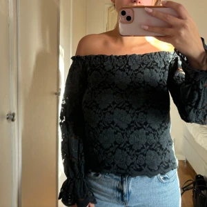 Svart offshoulder spetsblus från NA-KD - Säljer en superfin svart offshoulder blus från NA-KD i storlek 36. Den har ett vackert blommigt spetsmönster och långa ärmar med volangdetaljer vid ärmsluten. Perfekt för både fest och vardag! 🌸