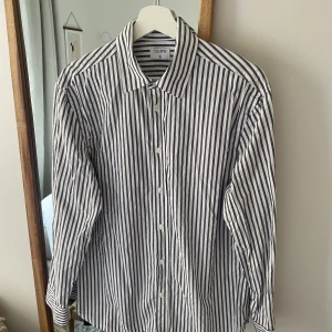 Randig skjorta från Filippa K - Säljer en stilren och klassisk randig skjorta från Filippa K i storlek 48. Skjortan är i bomull och har en snygg vit och blå färgkombination. Perfekt för både vardag och festliga tillfällen. Den har långa ärmar och knappar framtill. Skjortan är i mycket bra skick och gjord av ekologisk bomull.