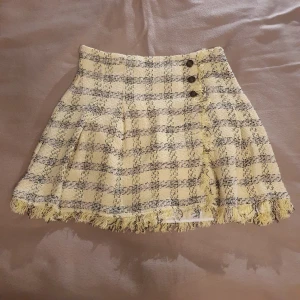 Rutig tweedkjol från Zara - Superfin rutig kjol från Zara i gult och svart. Den har en chic fransdetalj längst ner och tre dekorativa knappar framtill. Funkar både till vardags och fest. Kjolen är i mycket bra skick och har en bekväm passform. Foder/imnerkjol i bomull vilket gör den sååå bekväm! Material: 35% bomull, 65% polyester 