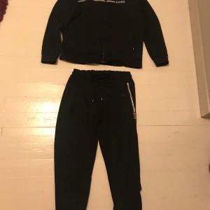 Svart träningsset från Boss - Säljer ett svart träningsset från Boss i mycket bra skick. Jackan har dragkedja och långa ärmar, medan byxorna har snörning i midjan. Perfekt för träning eller en avslappnad dag. Setet är stilrent med Boss-loggan på både jackan och byxorna.