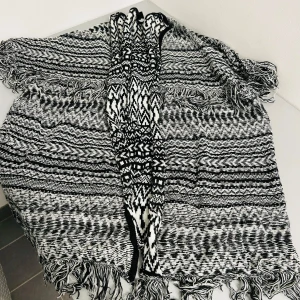 Svartvit mönstrad poncho från Lindex - Säljer en superfin svartvit poncho från Lindex i ett snyggt mönster. Den är stickad och har fransar i kanterna som ger en extra touch. Perfekt för att hålla sig varm under höst och vinter. Passar till alla outfits och är i bra skick!