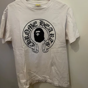Chrome x bape tee - Säljer en cool vit t-shirt från A Bathing Ape med tryck av Chrome Hearts och Baby Milo. T-shirten är i bomull och har korta ärmar. Perfekt för en avslappnad stil eller streetwear-look. Passar bra till jeans eller shorts!