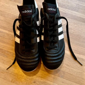 Adidas Copa Mundial Fotbollsskor - Säljer ett par klassiska Adidas Copa Mundial fotbollsskor i svart och vitt. Skorna är i nytt skick och har en snygg design med snörning och de ikoniska tre ränderna på sidan. Perfekta för fotbollsspelare som vill ha både stil och funktion på planen.
