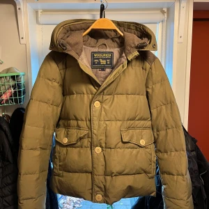 Olivgrön dunjacka från Woolrich - Säljer en olivgrön dunjacka från Woolrich i mycket bra skick. Jackan har en quiltad design med knappar och dragkedja framtill. Perfekt för höst, vinter och vår. Nypris på jackan är ca 6000, pris kan dock vara diskuterbart vid snabb affär😊