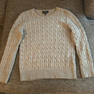 Kabelstickad tröja från Polo Ralph Lauren - Säljer en snygg grå kabelstickad tröja från Polo Ralph Lauren i storlek L(14-16). Tröjan har en klassisk design med långa ärmar och ribbade muddar. Perfekt för höst och vinter! Den har också den ikoniska broderade Ralph Lauren logotypen på bröstet. Passar perfekt för så väl finmiddag som skola. Tröjan har inga defekter och är väl omhändertagen! PRIS KAN DISKUTERAS VID SNABB AFFÄR!!