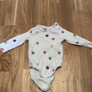 Vit prickig body från H&M - Söt vit body från H&M i ekologisk bomull med färgglada prickar i rosa, blått och brunt. Bodyn har långa ärmar och praktiska knappar i grenen för enkel påklädning. Perfekt för de små! 🌟