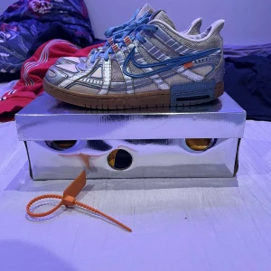 Nike off white  Rubber Dunk - Snygga Nike Air Rubber Dunk i vitt och blått med unika detaljer och snörning. Skorna har en robust sula och en modern design som sticker ut. Orginal låda, OW taggen ingår. Väl använda men har mer o ge, hål i hälarna men känns inte vid användning. Passar 44,5-45