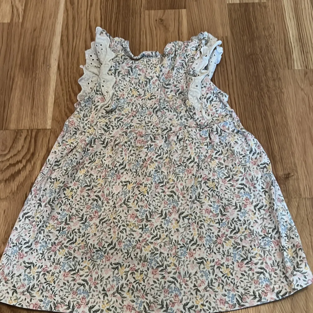 Söt blommig klänning från Newbie i storlek 74 för 6-9 månader. Klänningen har volangdetaljer och är ärmlös, perfekt för varma dagar. Den har ett fint blommönster i mjuka färger som rosa, blå och gul.. Mekot.