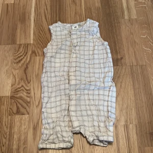 Vit ärmlös babyromper från H&M - Säljer en söt vit ärmlös babyromper från H&M med rutigt mönster. Den har knappar framtill för enkel påklädning. Perfekt för varma dagar!