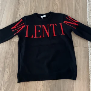 Valentino hoodie - Säljer en snygg svart tröja från Valentino med röd text över bröstet och ärmarna. Tröjan har en tight passform i storlek xl de vill säga passar M,L,XL och är perfekt för både höst och vinter. Den är i 10/10 skick och passar perfekt för en stilren look.
