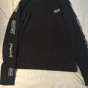Svart tröja från EA7 Emporio Armani - Säljer en svart tröja från EA7 Emporio Armani. Den har långa ärmar med coola logotyper längs sidorna och en liten logga på bröstet. Perfekt för en stilren och sportig look. Passar både till vardags och träning!