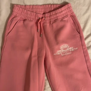 Rosa mjukisbyxor från Juicy Couture - Rosa mjukisbyxor från Juicy Couture med elastisk midja och snörning. Byxorna har ett broderat emblem på framsidan och texten 'In Juicy We Trust' på baksidan. Perfekta för en avslappnad stil.