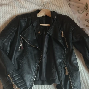 Svart skinnjacka från Voyelles - Snygg svart skinnjacka från Voyelles med dragkedjor och knappar. Jackan har en cool bikerstil med detaljer som bälten och quiltade partier på axlarna. Perfekt för en edgy look!