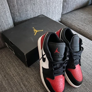Nike Air Jordan 1 Low strl 40 - Snygga Nike Air Jordan 1 Low i svart, vitt och rött. Skorna har en klassisk design med snörning och den ikoniska Jumpman-loggan på plösen. Perfekta för en stilren look. Använda 2 ggr. Nypris 1695:-