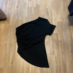 Svart topp - Elegant svart topp med asymmetrisk design och draperade detaljer. Perfekt för en stilren look. Helt oanvänd. 