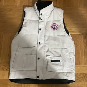 Vit dunväst från canada goose äkta Väst - Säljer en stilren vit dunväst från Canada Goose med knappar och en broderad logotyp på bröstet. Västen har praktiska fickor och är perfekt för kyligare väder. Den är tillverkad i Kanada och kombinerar funktion med mode.