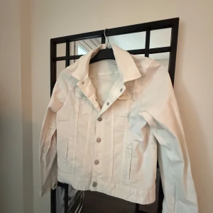 Vit jeansjacka - Snygg vit jeansjacka med knappar framtill och klassisk krage. Jackan har långa ärmar och två bröstfickor med knappstängning. Perfekt för en stilren look.