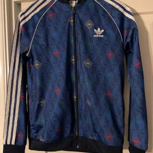 Blå mönstrad jacka från Adidas - Snygg blå jacka från Adidas med ett unikt mönster i olika färger. Jackan har dragkedja och de klassiska tre ränderna längs ärmarna. Perfekt för en sportig look.