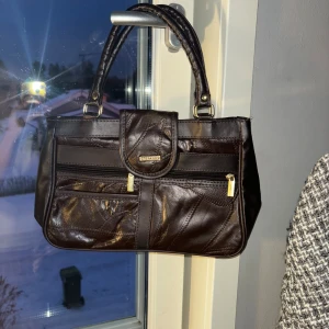 Brun handväska från Delmano - Snygg brun handväska från Delmano i skinn med praktiska dragkedjor och ett stilrent spänne framtill. Perfekt för att bära dina dagliga nödvändigheter med stil. Väskan har två robusta handtag för enkel bärning.