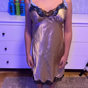 Champagnefärgad sidenklänning med spetsdetaljer - Elegant champagnefärgad klänning i siden med svarta spetsdetaljer vid halsringning och fåll. design med justerbara axelband för perfekt passform. Perfekt för en glamorös kväll.