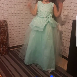 Mintgrön långklänning med volangdetaljer - Charmig mintgrön långklänning med volangdetaljer och en vacker rosett i midjan. Klänningen är ärmlös och har ett lager av tyll som ger en drömlik känsla. Perfekt för speciella tillfällen. För barn.