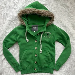 Tröja från Abercrombie & Fitch med päls - Snygg grön huvtröja från Abercrombie & Fitch med knappar och fuskpäls på huvan. Tröjan har ribbade muddar och en broderad logga på bröstet. Perfekt för kyliga dagar!💞🫶