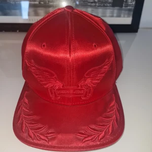 Röd keps från Robin's Jean - Snygg röd keps från amerikanska märket Robin's Jean. Köptes i New York och har aldrig blivit använd. Perfekt för att sticka ut i mängden och något utav en lyxigare keps. Reglerbar storlek. Garderobsrensning så kika gärna på övriga auktioner.