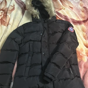Svart dunjacka från Canada Goose - Säljer en svart dunjacka från Canada Goose med pälsfodrad huva och broderad logotyp på ärmen. Jackan har dragkedja och knappar framtill för extra skydd mot kyla. Perfekt för kalla vinterdagar.