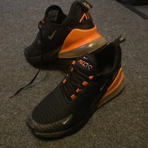 Nike Air 270 i svart och orange - Säljer ett par Nike Air 270 sneakers i svart med orange detaljer. Skorna har en modern design med synlig Air-enhet i hälen och snörning framtill. Perfekta för en sportig look.