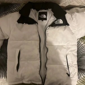 Vit och svart pufferjacka från The North Face - Säljer en snygg vit pufferjacka från The North Face med svart krage och dragkedja. Jackan har en klassisk design med två fickor framtill och The North Face-logga på bröstet och ryggen. Perfekt för kyliga dagar!