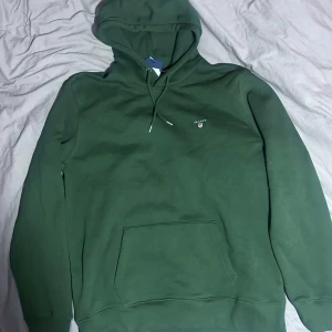 Grön hoodie från GANT - Säljer en snygg grön hoodie från GANT med broderad logga på bröstet. Den har en klassisk känguruficka och justerbar huva med snören. Perfekt för en avslappnad stil.