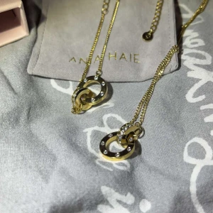 Guldhalsband och armband från Edblad  - Elegant guldhalsband samt tillhörande armband från Edblad med två sammanlänkade ringar, varav en är dekorerad med gnistrande stenar. Perfekt för att ge en touch av glamour till din outfit. Priset är för båda tillsammans.
