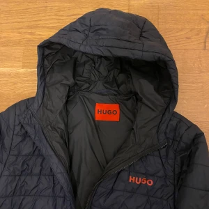Svart quiltad jacka från Hugo - Snygg svart quiltad jacka från Hugo med huva och dragkedja. Perfekt för kyligare dagar. Jackan har en stilren design med märkets logga i rött på bröstet.  Och använd knappt men de finns bara ett litet hål där ni ser och bakom bara en jätte liten scratch 