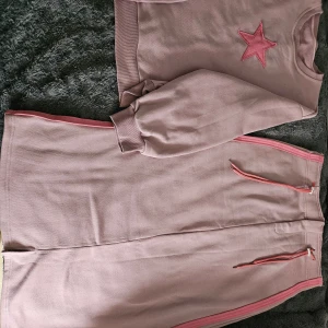 Rosa set med stjärndetalj - Säljer ett mysigt rosa set med en långärmad tröja och matchande byxor. Tröjan har en stjärndetalj på bröstet och byxorna har rosa dragkedjor. Perfekt för en avslappnad dag hemma eller ute.