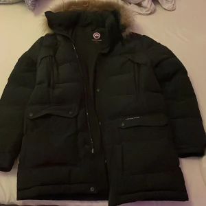 Svart dunjacka från Canada Goose - Säljer en svart dunjacka från Canada Goose med pälskantad huva. Jackan har en dragkedja och knappar framtill samt flera praktiska fickor. Perfekt för kalla vinterdagar. Säljer den billigt pga av den defekten på bild 3 men kan sy ihop den innan den skickas iväg tar inte så lång tid:)