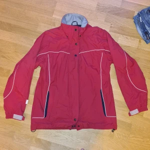 Röd vindjacka med dragkedja - Vindjacka i okänt märke. Den har flera fickor. Fin och hel men väl använd.