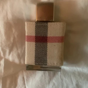 Burberry London Parfym - Elegant parfym från Burberry med klassiskt rutmönster i rött, svart och beige. Flaskan är klädd i tyg och har en stilren design. Perfekt för den som älskar tidlös brittisk stil. Använd 2-3 gånger,