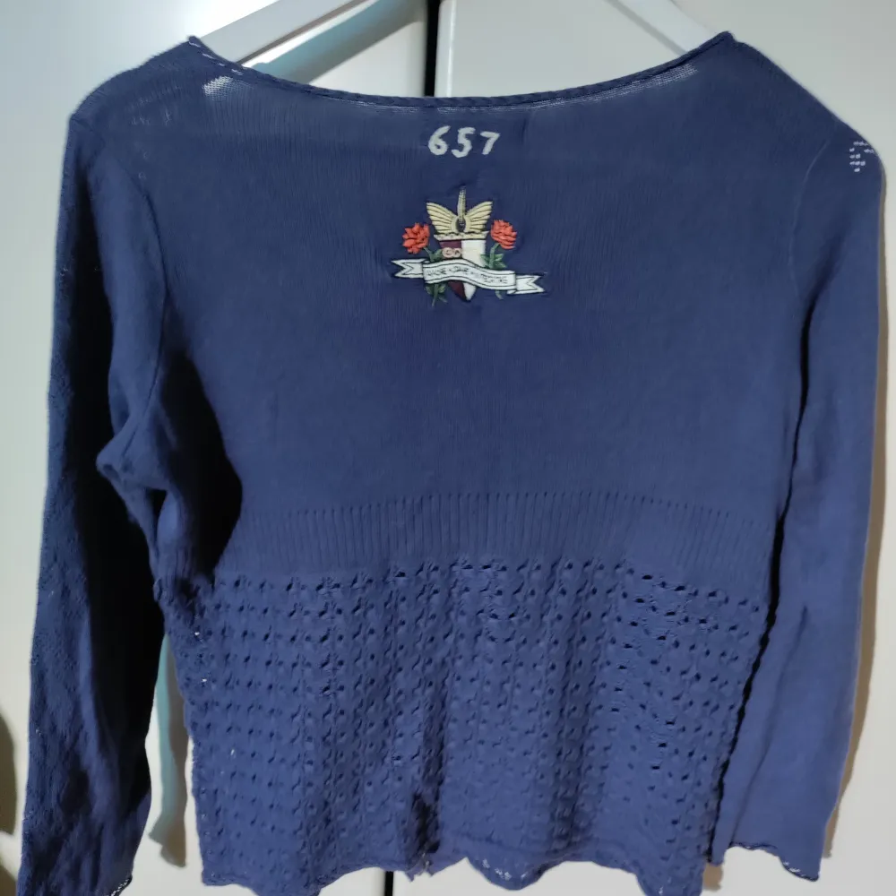 Säljer en mörkblå kofta med vackert broderi på ryggen. Koftan har långa ärmar och knappar framtill. Perfekt för en stilren look.. Neuletakit & Villapaidat.