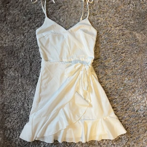 Vit omlottklänning från Parisian - Säljer en söt vit omlottklänning från Parisian med volangdetaljer och knytband i midjan. Klänningen har smala axelband och en v-ringad halslinje, perfekt för sommarens festligheter.