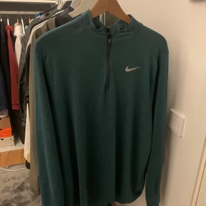 Grön långärmad tröja från Nike - Snygg grön långärmad tröja från Nike med dragkedja vid halsen och det klassiska Nike-loggan på bröstet. Perfekt för träning eller i vardagen. Tröjan har en stilren design och är gjord i ett bekvämt material.
