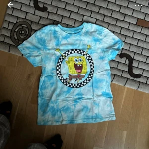 Blå VANS tie-dye t-shirt med Svampbob - Cool blå tie-dye t-shirt med en stor bild av Svampbob Fyrkant i mitten. Perfekt för fans av den tecknade serien! T-shirten har korta ärmar och en rund halsringning.