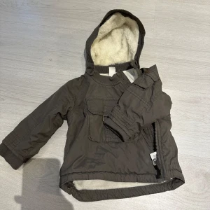 Brun jacka med fleecefoder - Säljer en brun jacka med mysigt fleecefoder och huva. Jackan har en praktisk dragkedja på sidan och en ficka framtill. Perfekt för kyliga dagar! Perfekt jackan för barn 6-12 månader