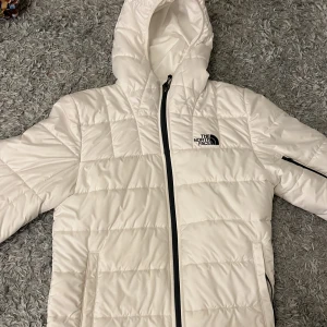 Vit dunjacka från The North Face - Snygg och varm vit dunjacka från The North Face med huva och dragkedja. Jackan har en quiltad design och praktiska fickor med dragkedja. Perfekt för kyliga dagar.knappt använt.
