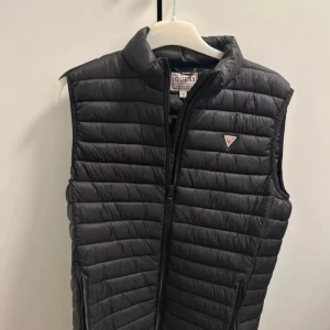 Svart dunväst från Guess - Snygg svart dunväst från Guess med dragkedja och det klassiska Guess-emblemet på bröstet. Perfekt för lager-på-lager under kyliga dagar. Västen har en stilren design och är lätt att matcha med olika outfits. Denna väst är mycket sällsynt.