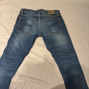 Blå Nudie Jeans - Säljer ett par snygga blå jeans från Nudie Jeans i modellen Tight Terry. De är tillverkade av 99% ekologisk bomull och 1% elastan för en bekväm passform. Jeansen har en midwaist och en tight stil med klassiska femficksdetaljer och orange sömmar. Passar inte mig därav priset.