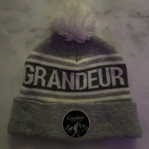 Grå mössa med tofs från Grandeur - Snygg grå mössa från Grandeur med vit tofs och text. Den har ett stilrent randigt mönster och en uppvikt kant med en rund logga framtill. Perfekt för kyliga dagar!